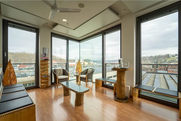 Wartebereich mit Rattansesseln, Steintisch und Stehtisch vor bodentiefen Fenstern mit Blick über Passau
