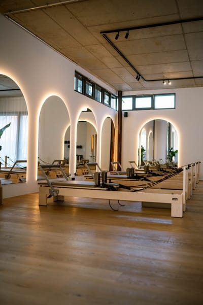 Reihe von Pilates-Reformern aus hellem Holz vor beleuchteten, bogenförmigen Spiegeln